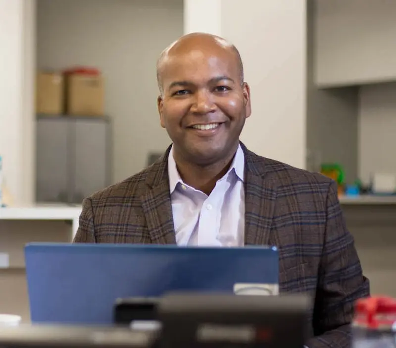 A man in business casual sitting at a desk and smiling